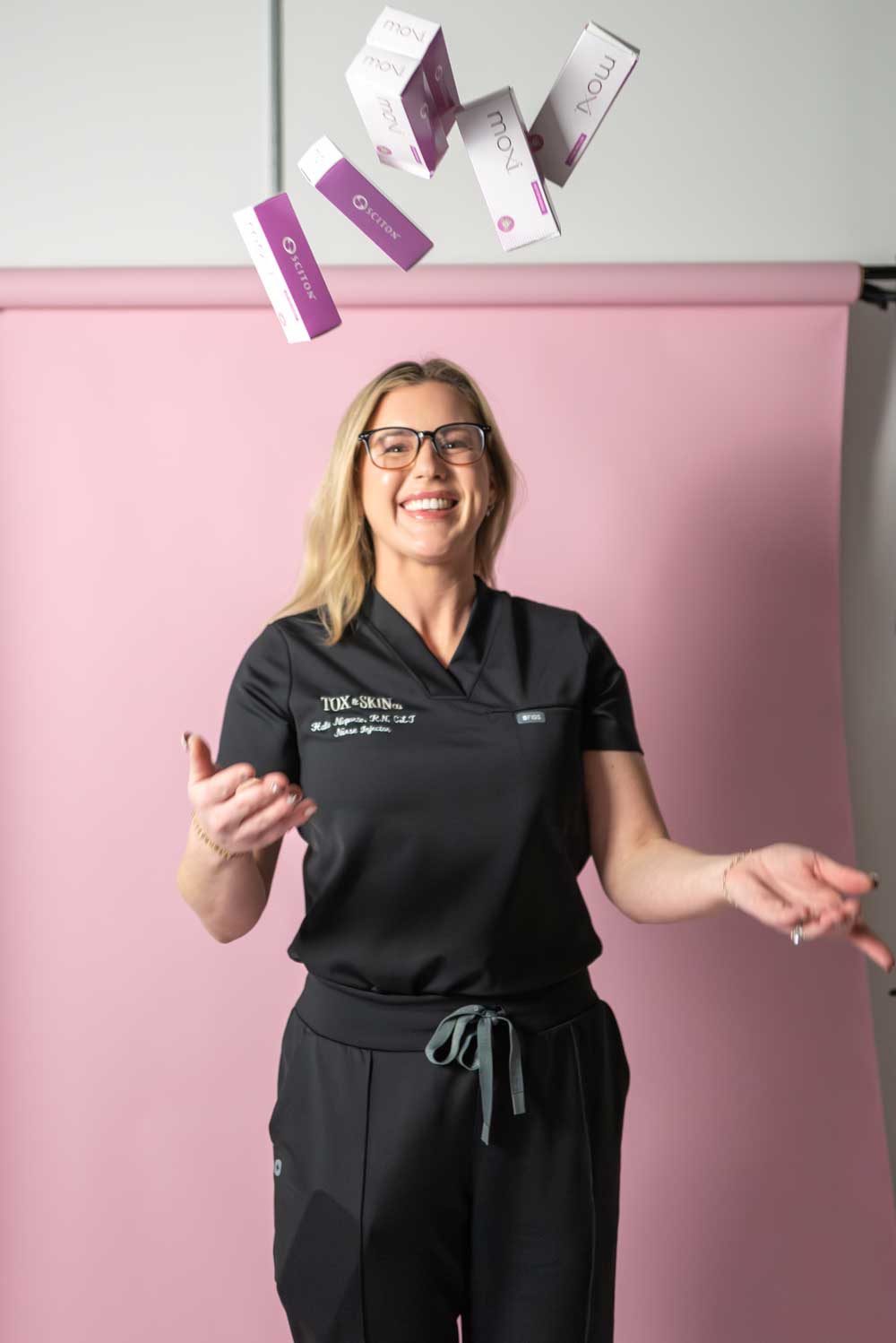 A woman in black scrubs smiles while tossing skincare product boxes in the air, against a pink background.